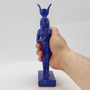 Op de afbeelding: Een blauw lapis lazuli beeld van een Egyptische godin met een kroon en een staf. Het beeld staat op een rechthoekige basis.