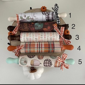 May include: Five mini rolling pins with fall-themed designs. The rolling pins are decorated with plaid, pumpkins, and fall-themed text. The rolling pins are arranged vertically on a wooden surface.