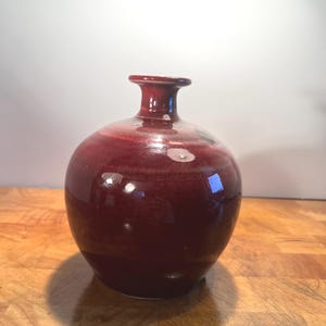 May include: A round, maroon-colored ceramic vase with a narrow neck. The vase has a glossy finish and reflects light. The vase is a decorative item for home decor.