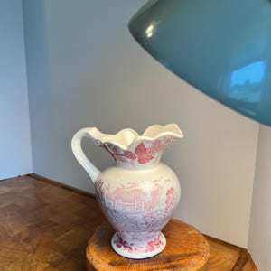May include: A white and pink ceramic pitcher with a detailed landscape design. The pitcher has a ruffled rim and a curved handle. It sits on a small, round wooden stand.