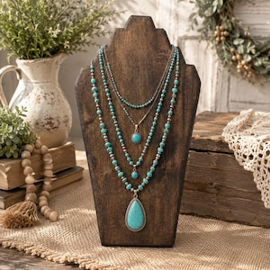 May include: A multi-strand necklace featuring turquoise-colored beads and silver-toned metal accents. The design includes three distinct layers, with two small oval turquoise pendants and one large, prominent teardrop-shaped turquoise pendant.