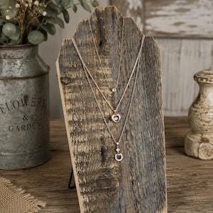 May include: A rustic wooden jewelry display stand featuring a weathered, textured grain, holding three layered necklaces. The top necklace has a small clear stone, the middle has a heart-shaped pendant, and the bottom has a horseshoe-shaped charm.