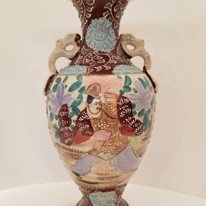 May include: An elaborate ceramic vase with a cream-coloured rim and base. The vase has a maroon neck and base with teal and white floral and geometric patterns. The body of the vase depicts a figure in traditional attire, with dragon-shaped handles.