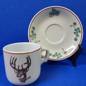 May include: A white ceramic coffee cup with a brown rim and a brown and white image of a deer's head with antlers. The cup is paired with a white ceramic saucer with a brown rim and a green and brown design of pine branches and cones.