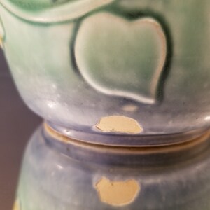 May include: A blue and green ceramic vase with a leafy design. The vase has a chip on the bottom edge.