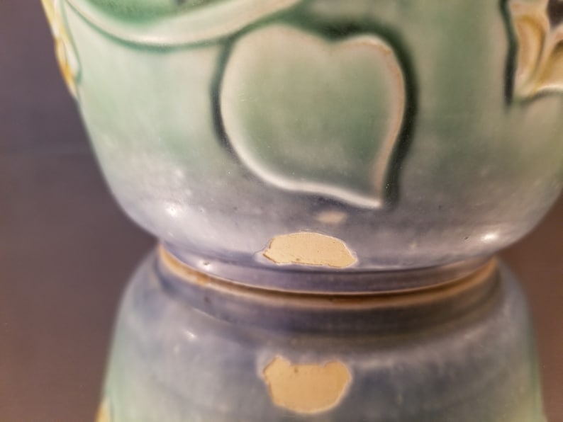 May include: A blue and green ceramic vase with a leafy design. The vase has a chip on the bottom edge.