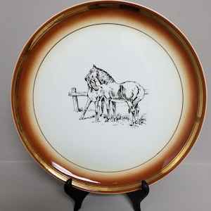 Puede incluir: Un plato de cerámica blanco con un borde marrón y un ribete dorado presenta una ilustración en blanco y negro de dos caballos. El plato está sobre un soporte negro.
