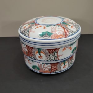 Op de afbeelding: Een ronde keramische container met twee niveaus en een deksel. De container is wit met een blauwe rand en is versierd met een kleurrijk, gedetailleerd Aziatisch geïnspireerd ontwerp met bomen, vogels en andere natuurlijke elementen. Het deksel heeft een klein, cirkelvormig ontwerp.