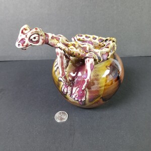 Puede incluir: Una figurita de dragón de cerámica con un esmalte blanco, rosa y marrón. El dragón está posado sobre una base redonda de cerámica marrón con un esmalte rosa y dorado.