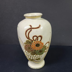 May include: A small, white ceramic vase with a crackled glaze. It features a floral design with orange, red, green, and gold accents. The vase has a rounded body and a short, flared base.