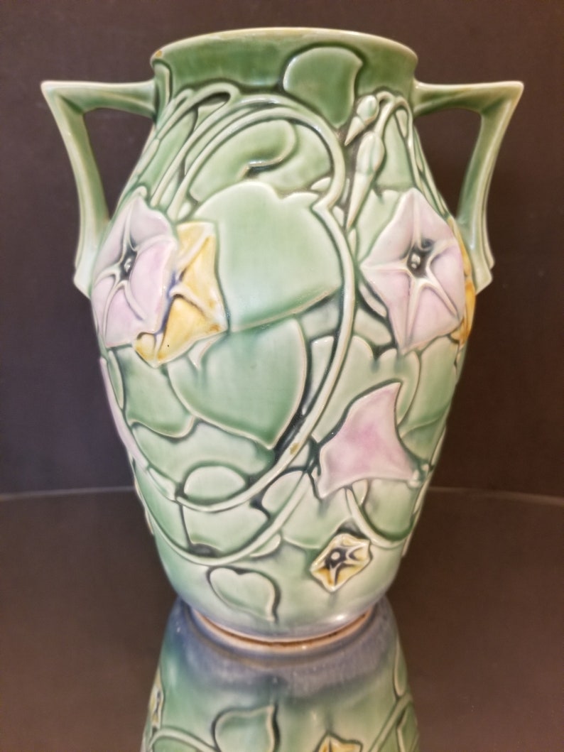 May include: A green ceramic vase with two handles and a floral design. The vase is decorated with pink and yellow flowers and green leaves.