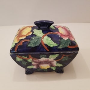 Puede incluir: Una caja de cerámica azul con un diseño floral. La caja tiene una tapa con un asa elevada y está decorada con flores en tonos rosa, amarillo y verde.