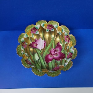 May include: A decorative bowl with a scalloped edge and a gold background. The bowl is decorated with a floral design featuring pink and purple irises, green leaves, and yellow flowers.