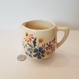 Pode incluir: Um pequeno creamer de cerâmica de cor creme com um design floral. As flores são pintadas em tons de azul, amarelo, laranja e vermelho.