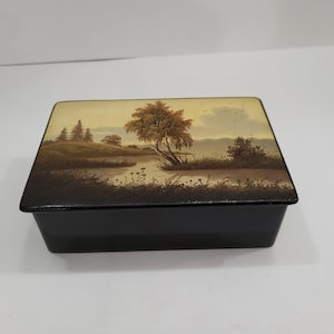 May include: A rectangular black box with a hinged lid featuring a painted landscape scene. The artwork depicts a calm body of water, grassy banks, and trees in shades of brown, tan, and muted green under a soft, light-coloured sky.