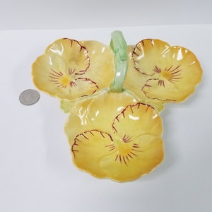 May include: A yellow ceramic serving dish shaped like three pansies with a green handle. The pansies have red and yellow details.