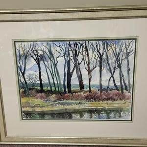 Puede incluir: Pintura a la acuarela de un paisaje con árboles y una masa de agua. La obra de arte presenta un marco plateado y un passe-partout blanco. Los árboles están representados con troncos y ramas oscuras, con una mezcla de follaje verde, morado y marrón.