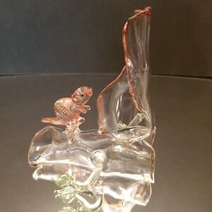 May include: A glass sculpture of a small animal, possibly a squirrel, perched on a branch. The sculpture is made of clear glass with a pink tint and features a green leaf.