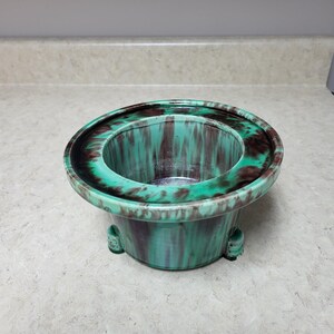 Peut inclure: Un pot de fleurs rond en céramique avec un vernis brillant présentant un motif marbré de brun foncé et de vert turquoise vif. Le récipient possède un large bord plat et repose sur trois petits pieds intégrés à la base, créant un profil bas.