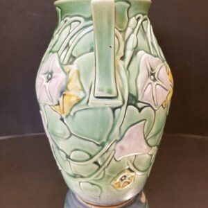 May include: A green ceramic vase with two handles and a floral design. The vase is decorated with pink and yellow flowers and green leaves.