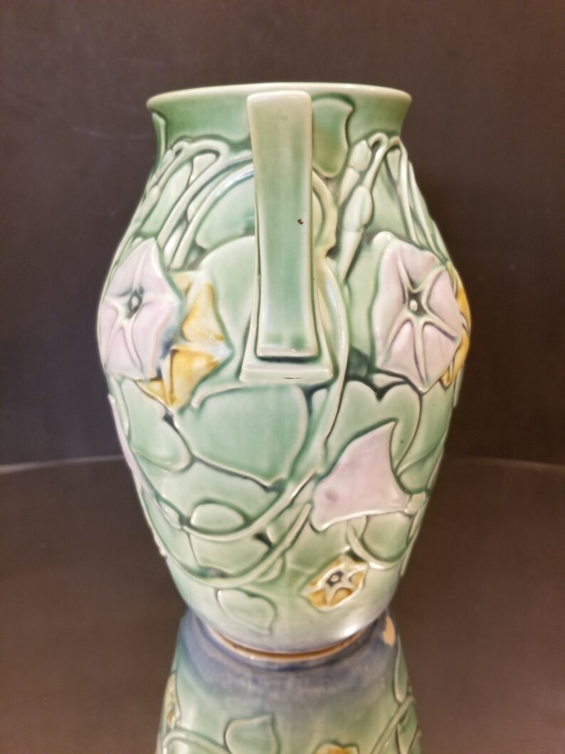 May include: A green ceramic vase with two handles and a floral design. The vase is decorated with pink and yellow flowers and green leaves.