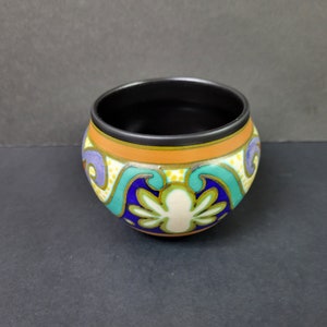 May include: A small, round ceramic bowl with a black interior and a colourful, geometric design. The design features swirls of blue, green, orange, and white on a cream background.
