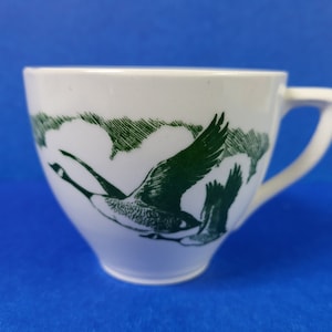 Puede incluir: Taza de té de cerámica blanca con un diseño verde de dos gansos volando en el cielo. La taza tiene una forma redondeada y un asa.