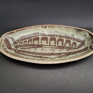 May include: An oval-shaped ceramic dish featuring a dark brown base with a series of repeating rectangular cutouts along the centre. The surface is finished with a mottled, light green and cream-coloured glaze that drips over the brown edges.