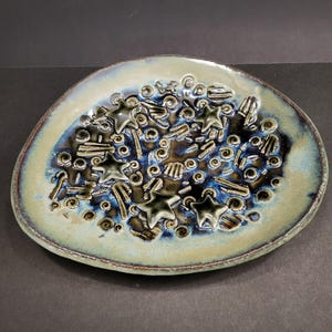 May include: An oval ceramic dish featuring a textured surface with raised star shapes and swirling patterns. The glaze transitions from a light beige rim to a deep blue and black centre, creating a contrast between the smooth edge and the detailed interior.