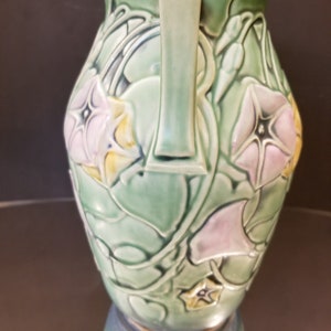 May include: A green ceramic vase with two handles and a floral design. The vase is decorated with pink and yellow flowers and green leaves.