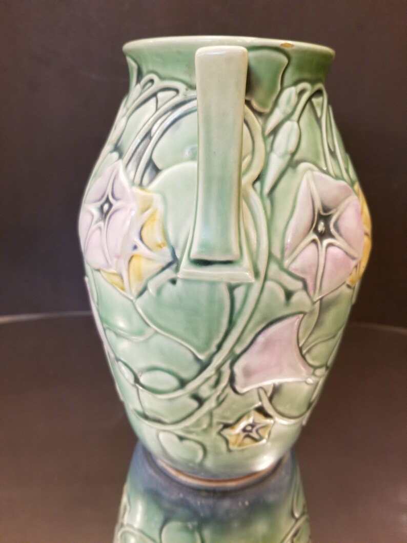 May include: A green ceramic vase with two handles and a floral design. The vase is decorated with pink and yellow flowers and green leaves.
