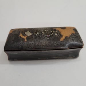 Pode incluir: Uma caixa retangular de madeira escura com uma superfície texturizada e um design floral decorativo. A tampa apresenta um padrão castanho claro e preto com detalhes prateados e rosa. A caixa tem cerca de 10 cm de comprimento.