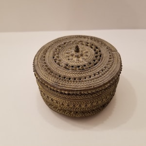 Op de afbeelding: Een ronde, sierlijke metalen doos met een deksel. De doos is versierd met ingewikkelde patronen en heeft een klein knopje bovenop.