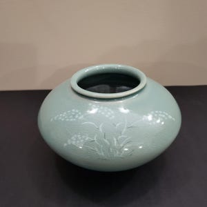 以下が含まれることがあります： 丸い、薄緑色の陶器の花瓶。広い開口部があり、表面には白い花と葉の模様が描かれています。花瓶はひび割れた釉薬で、暗い表面に置かれています。家庭用の装飾品です。