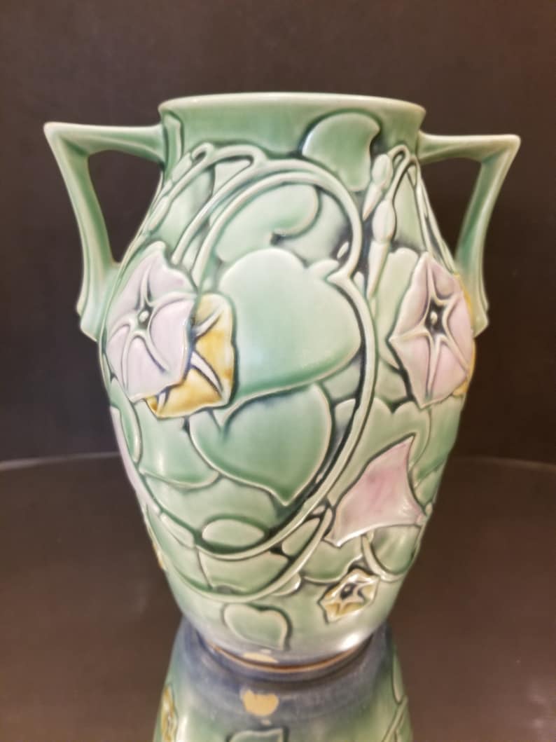May include: A green ceramic vase with two handles and a floral design. The vase is decorated with pink and yellow flowers and green leaves.