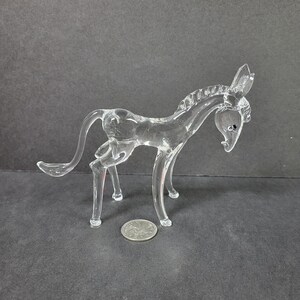 Puede incluir: Una figurita de caballo de vidrio transparente con una crin y una cola detalladas. El caballo está de pie sobre sus cuatro patas y tiene la cabeza baja. La figurita mide aproximadamente 10 cm de alto.