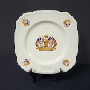 Może przedstawiać: Biały ceramiczny talerz z pozłacanym rantem i złotym wzorem w środku. Wzór przedstawia portret króla Jerzego VI i królowej Elżbiety, z tekstem "Long May They Reign". Talerz ma również pozłacaną obwódkę z tekstem "Compliments of The S.E. Gust Stores".