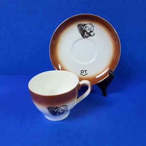 May include: A ceramic teacup and saucer set. The cup and saucer have a white interior and a brown exterior with a dog illustration. The saucer has a brown rim and a dog illustration. The cup has the letters "OT" on the side.