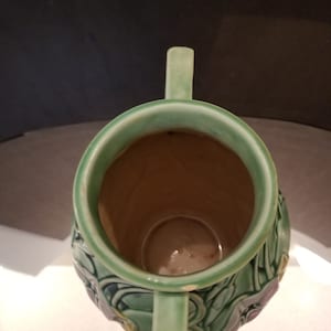 May include: A green ceramic vase with two handles and a floral design. The vase has a wide mouth and a narrow base.
