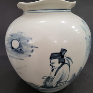 Pode incluir: Um vaso de cerâmica branca com borda recortada e ilustrações pintadas à mão em azul. O design apresenta uma figura sentada com vestes tradicionais, uma lua circular e marcas caligráficas ténues no corpo arredondado do vaso.