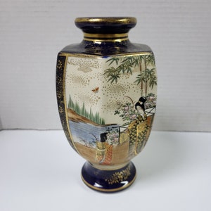 Puede incluir: Un pequeño jarrón de cerámica azul y dorado con forma hexagonal. El jarrón está decorado con una escena de una mujer con un kimono japonés tradicional de pie en un jardín con árboles de bambú y flores. El jarrón tiene un borde dorado y una base dorada.