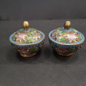 Könnte beinhalten: Zwei kleine, runde Schalen mit Deckel, die mit aufwendiger Cloisonné-Emaille-Arbeit verziert sind. Die Schalen zeigen ein florales Muster in Blau-, Grün-, Rosa- und Goldtönen mit einem goldfarbenen Rand und einem kleinen, strukturierten Goldknopf.