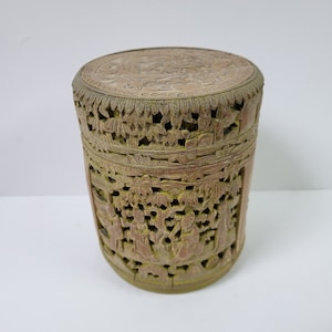 Puede incluir: Una caja redonda de madera tallada con diseños intrincados. La caja está hecha de madera marrón claro y tiene un patrón detallado de flores y hojas.