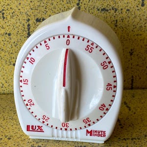 Könnte beinhalten: Weißer Küchentimer mit rotem Zifferblatt und Markierungen. Der Timer hat ein geschwungenes Design mit einem großen Zifferblatt in der Mitte, das mit Zahlen und roten Indikatoren markiert ist. Der Timer liest "LUX Minute Minder".
