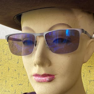 May include: A pair of rectangular spectacles with a matte grey metal frame and blue-tinted lenses. The spectacles feature a thin bridge and slim temples, resting on a mannequin head wearing a wide-brimmed brown hat and a dark purple shirt.