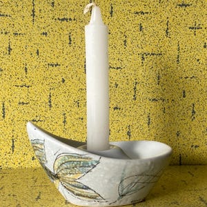 Pode incluir: Uma vela branca em um castiçal de cerâmica com um desenho de folhas. O castiçal é branco com desenhos de folhas verdes e amarelas. O fundo é uma superfície amarela com um padrão preto.