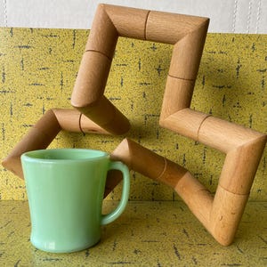 May include: A light green mug with a handle sits in front of a wooden geometric sculpture. The mug has a smooth, rounded shape. The sculpture is made of light brown wood and has a zig-zag design.