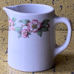 May include: White ceramic jug with a handle and spout. The jug is decorated with a floral design featuring pink flowers and green leaves. It is likely used for serving beverages.