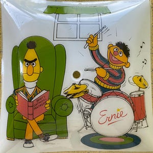 Puede incluir: Plato decorativo cuadrado con los personajes de Barrio Sésamo, Epi y Blas. Epi está sentado en un sillón verde leyendo un libro. Blas toca la batería, con el nombre "Ernie" en el bombo. Ilustración de una ventana.