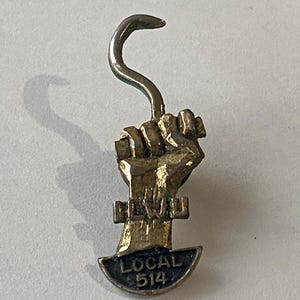 Könnte beinhalten: Vintage Metall-Anstecknadel mit einer geballten Faust, die einen Haken hält. Die Anstecknadel ist goldfarben und trägt die Aufschrift "IBEW" und "LOCAL 514". Der Haken ist oben gebogen.